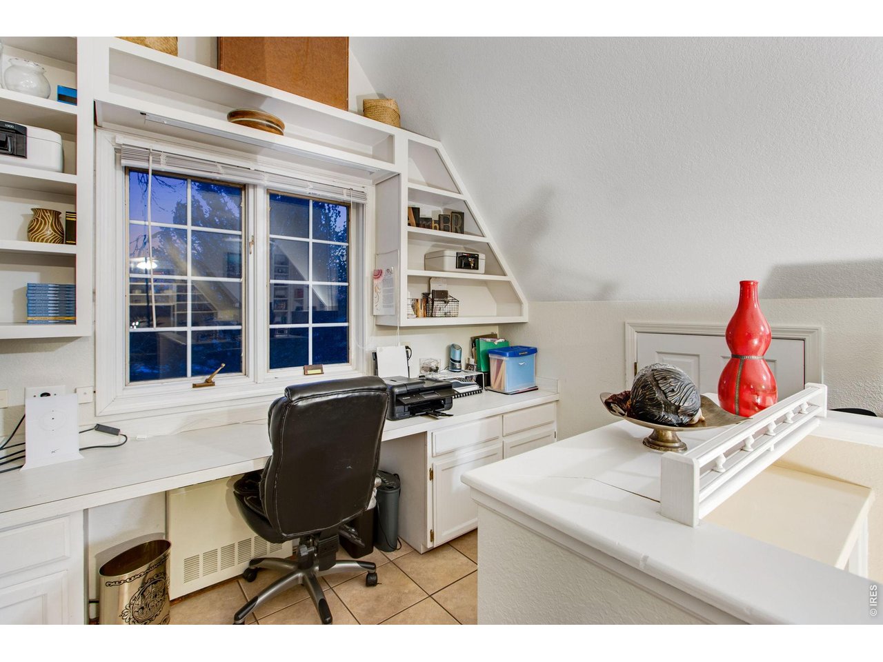 Office workstation with tile flooring, built-in wrap-around desk facing out the window. Built in shelving around the desk and bright overhead light. 